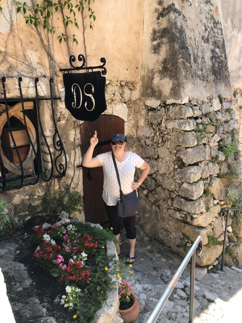 Darcy standing under a sign surrounded by stones walls and flowers pointing to a sign with the initials "DS" and smiling.