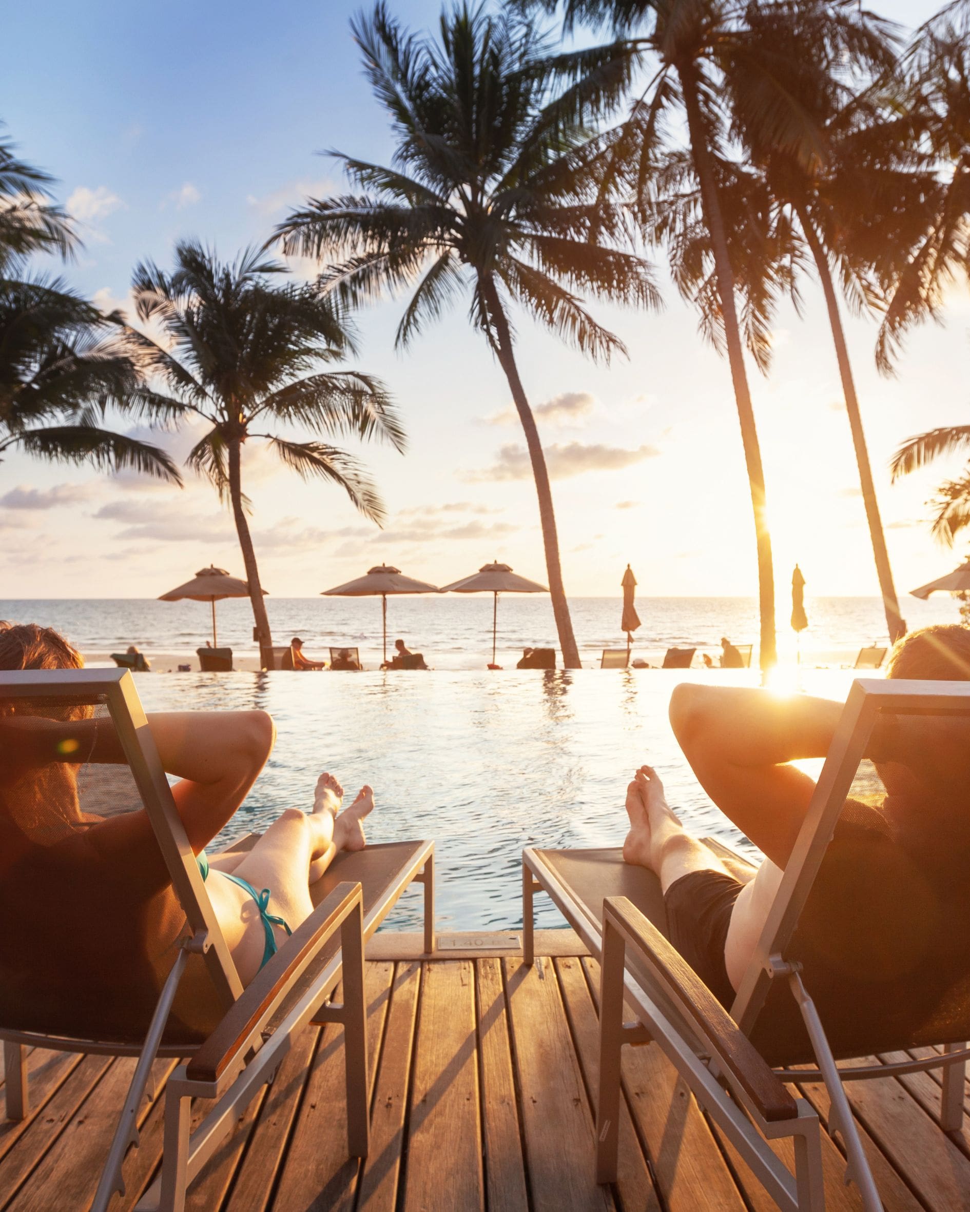 Two people relaxing on lounge chairs beside an infinity pool at a tropical beach resort during sunset, with palm trees, ocean views, and beach umbrellas in the background.
