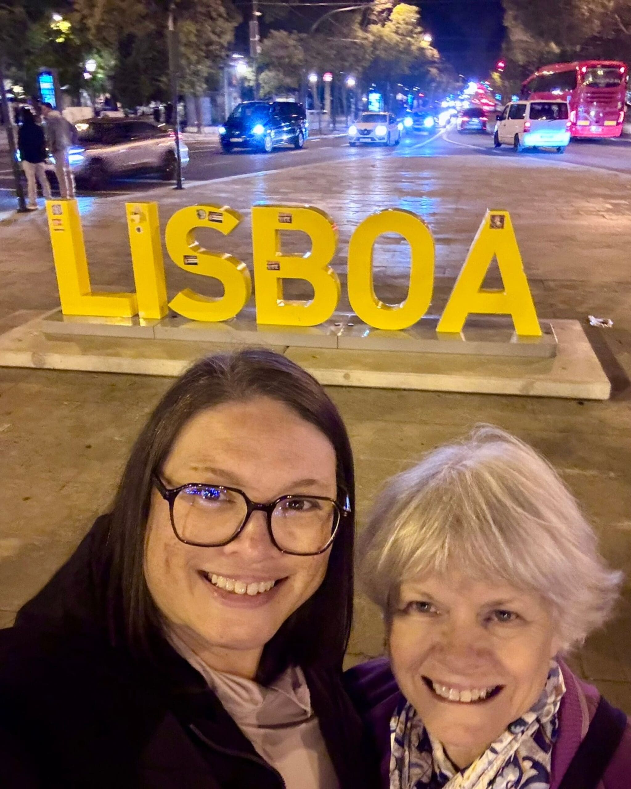 Two women smiling for a nighttime selfie in front of large yellow “LISBOA” letters on a city street in Lisbon, with traffic and streetlights in the background.