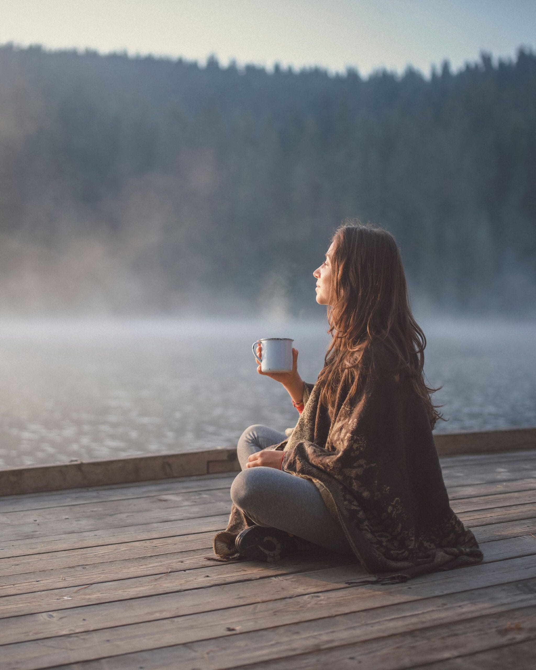 Woman sitting on a wooden dock beside a misty lake, holding a mug and reflecting quietly, representing solo travel, slow travel, and thoughtful travel experiences.
