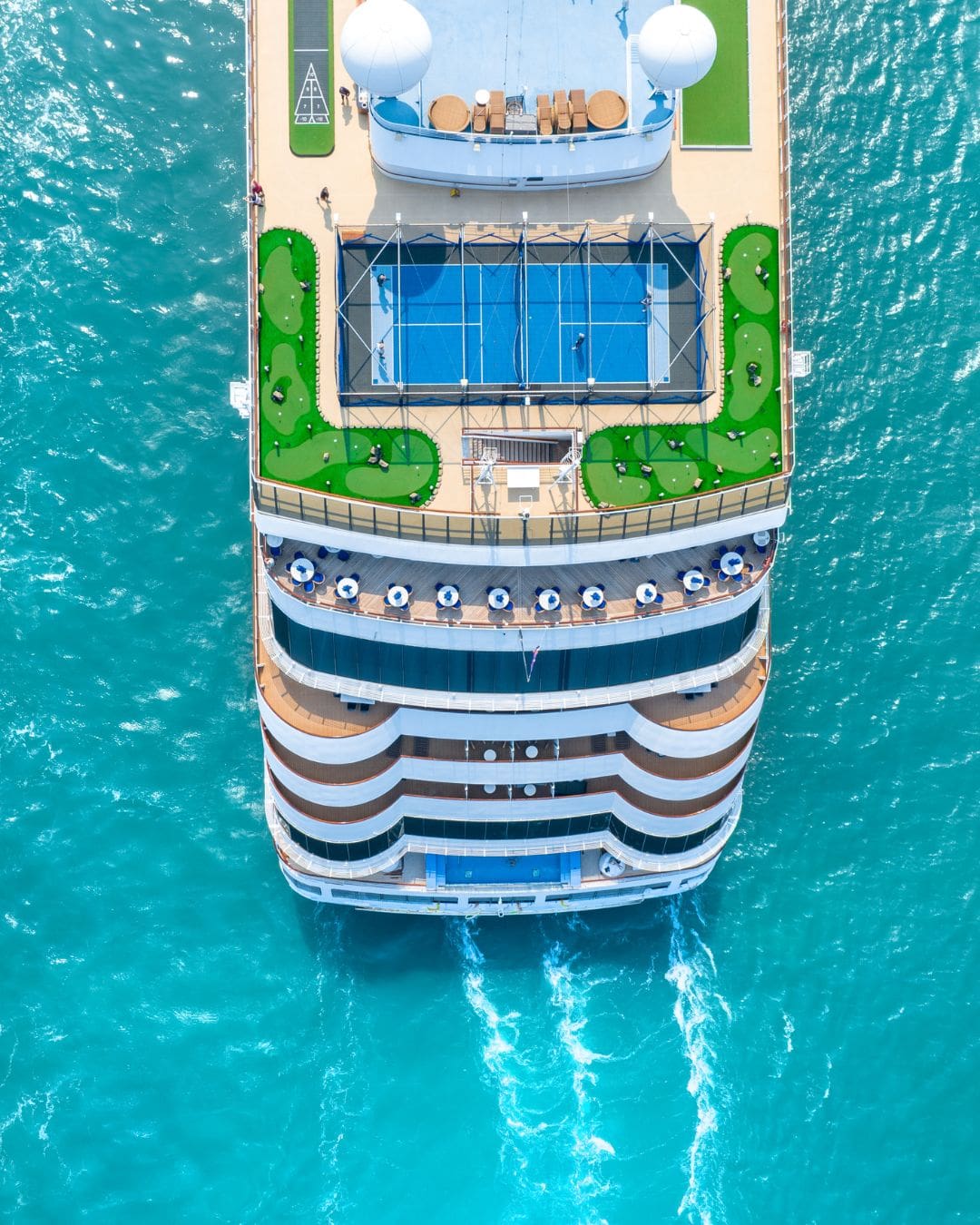 Aerial view of a modern cruise ship sailing through turquoise ocean waters, showing the top deck with a sports court, lounge seating, and open-air amenities.