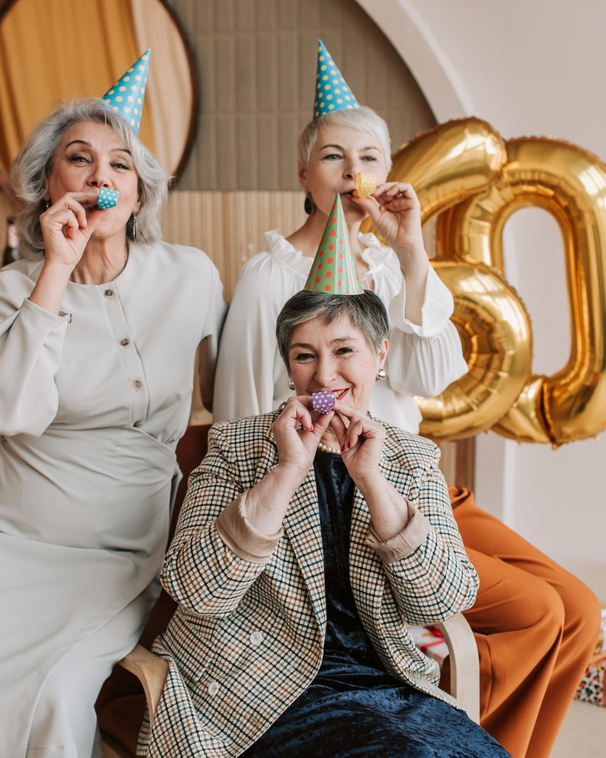 Three women celebrating a milestone birthday with party hats and noisemakers, symbolizing joyful milestone celebrations and meaningful moments shared together.