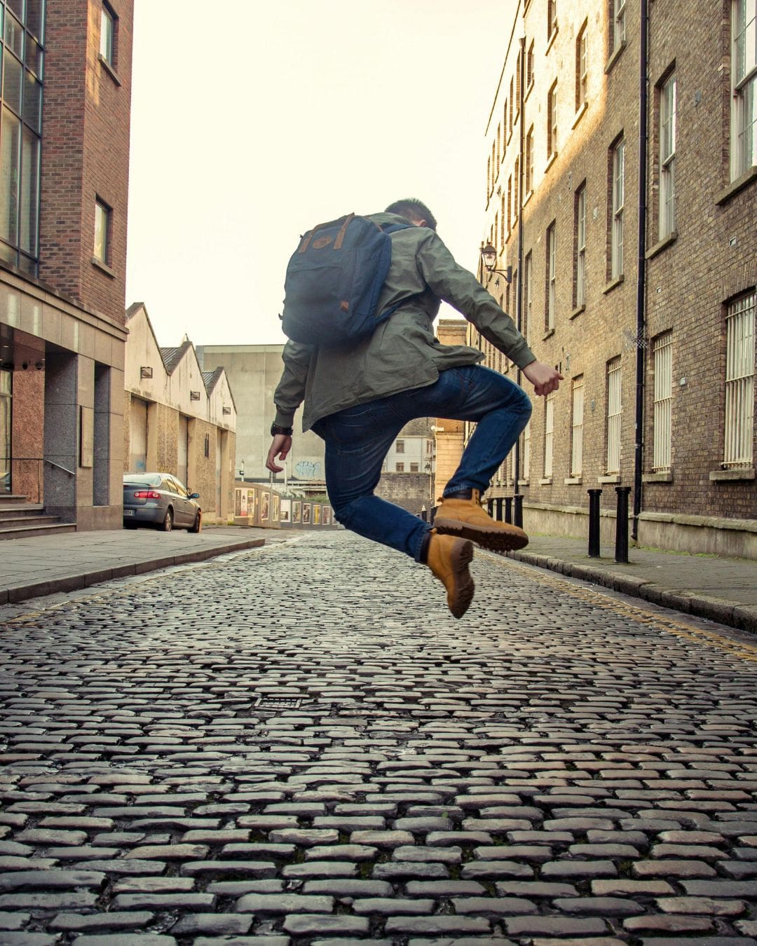 Traveler mid-jump on a cobblestone street in a European-style city, wearing a backpack and boots, expressing movement and spontaneity.