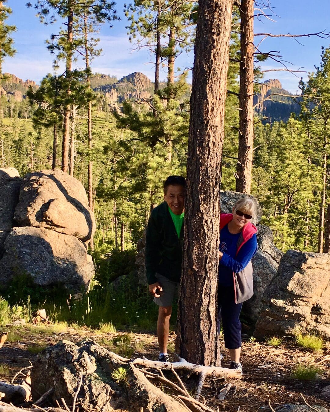Two travelers playfully peeking around a tree while hiking in the mountains.